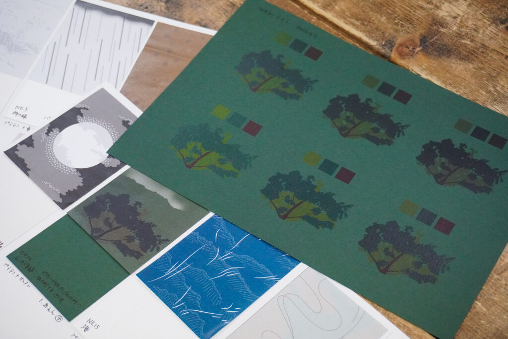 木製のテーブルの上に、「山と霧」のグラフィックに描かれた木々を、オンデマンド印刷により異なる濃度パターンで印刷した出力紙が置かれている。出力紙の下には『雨』の各グラフィックのサンプルが置かれている。