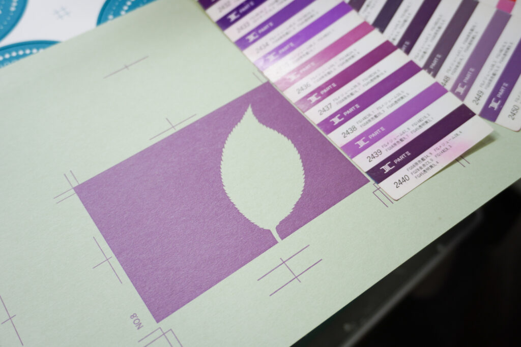 「葉っぱ」のグラフィックの紫色の背景ベタ面を印刷した出力紙が置かれている。印刷面の右横には指定色のカラーチップが置かれている。