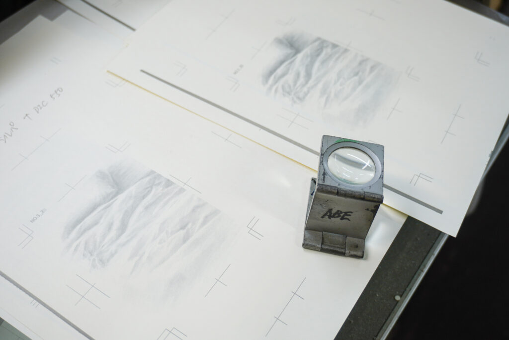 「二度寝」のグラフィックを出力した紙が複数枚重なっている。その上に版の刷り上がりを拡大して確認するためのルーペが置いてある。