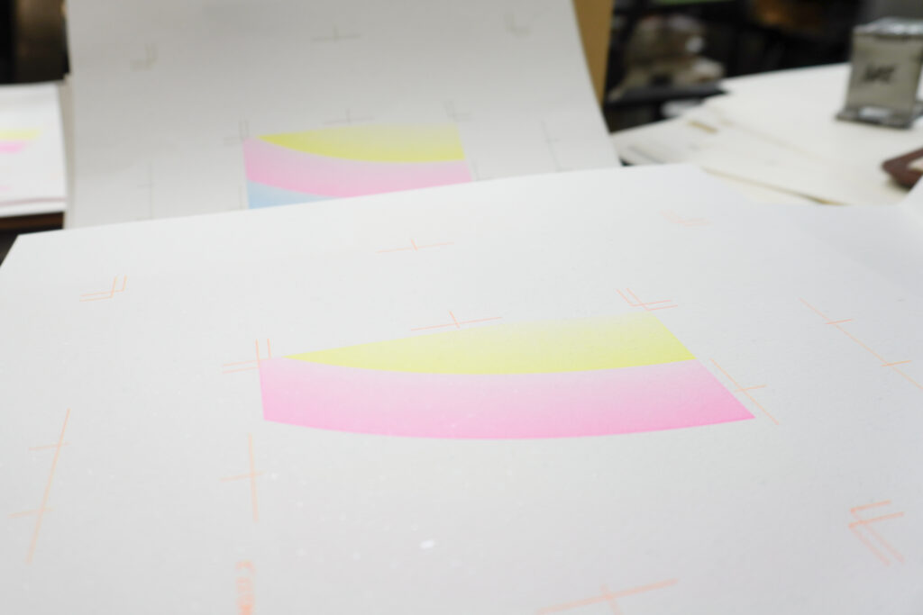 「虹」の出力紙を写した写真。手前に黄色とピンクの色面のみが刷られたもの、奥に黄色とピンク、水色の3色の色面が刷られたものが覗く。